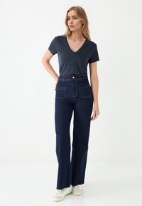 Czarna szara koszulka z dekoltem w serek zestawiona z wysokimi, ciemnoniebieskimi dżinsami z dwiema przednimi kieszeniami i żółtym przeszyciem, noszona z białymi trampkami.