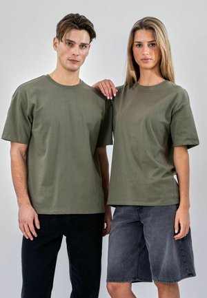 Mand og kvinde står side om side iført olivengrønne T-shirts; manden har sorte bukser på, kvinden har en grå denims nederdel, kvindens hånd hviler på mandens skulder.