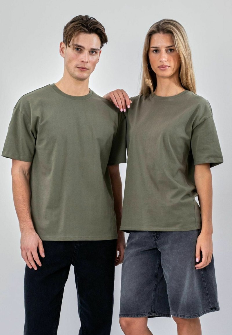 Un homme et une femme se tiennent côte à côte, portant des T-shirts vert olive ; l’homme porte un pantalon noir, la femme une jupe en jean grise, la main de la femme repose sur l’épaule de l’homme.