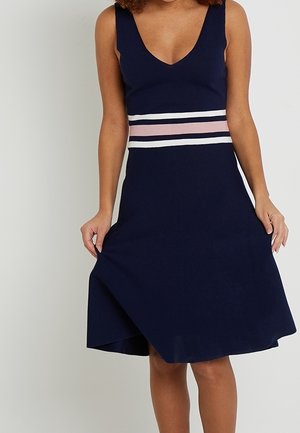 Femme portant une robe bleu marine sans manches avec un décolleté en V profond et des rayures horizontales roses et blanches à la taille, tenant l'ourlet de la robe.