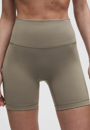 Vrouw draagt hooggetailleerde taupekleurige nauwsluitende fietsshorts, getoond van taille tot halverwege de dij tegen een effen achtergrond.