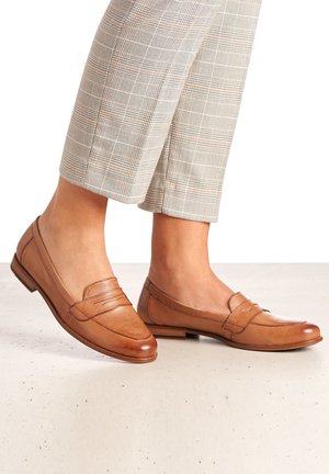 Pieds chaussés de mocassins en cuir beige assortis à un pantalon court écossais beige, debout sur une surface blanche.