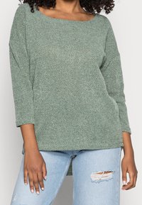 Grünes strukturiertes Strickoberteil mit weitem Boatneck-Ausschnitt und Dreiviertelärmeln. Kombiniert mit hellblauen, zerrissenen Jeans für einen Kontrast.