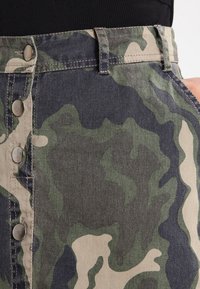 Falda de mezclilla con estampado de camuflaje que presenta una cintura ajustada, botones en tono plateado y una mezcla de colores verde, azul marino y beige.