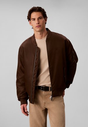Giovane uomo che indossa una giacca bomber marrone scuro sopra una camicia beige e pantaloni beige con cintura nera, su uno sfondo grigio chiaro uniforme.