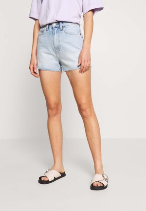 Persona que lleva pantalones cortos de denim claro de talle alto, camiseta de manga corta color lavanda y sandalias beige con hebillas metálicas circulares grandes.