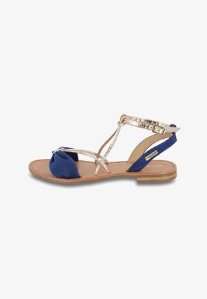 Sandal en azul marino con un frente anudado, tiras metálicas doradas y una suela plana en tono marrón. Presenta tiras cruzadas y una hebilla para el cierre.