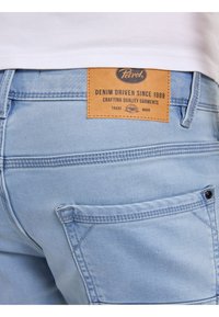 Los pantalones cortos de mezclilla de color azul claro presentan un parche de cuero de la marca con texto, un bolsillo lateral con botón y detalles de costura.