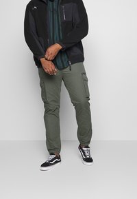 Hombre con chaqueta negra con cremallera, camiseta a rayas verdes, pantalones cargo oliva y zapatillas negras y blancas de pie contra un fondo liso.