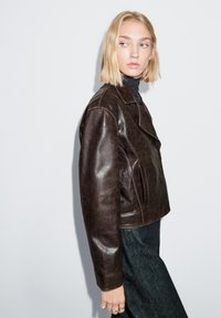 Braune Lederjacke mit kurzem Schnitt, Reverskragen und sichtbaren Nähten. Kombiniert mit einem schwarzen Rollkragenpullover und dunkler Denim.