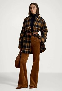 Γυναίκα που φοράει ένα καρό παλτό σε καφέ και μαύρο χρώμα με ζωνάκι, καφέ φαρδιές παντελόνες, μαύρο ζιβάγκο και κρατά μια καφέ στρογγυλή τσάντα.