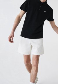 Polo noir à manches courtes avec col, arborant un petit logo vert. Associé à un short blanc et des baskets blanches.