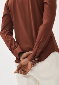 Roestkleurig overhemd met lange mouwen en knoopsluitingen, gladde stof, en een zijsplit aan de onderkant, gecombineerd met lichtbeige broek.
