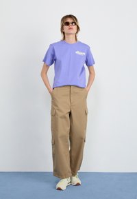 Purple cotton t-shirt with logo, paired with beige cargo pants featuring side pockets, worn with white and green sneakers.
