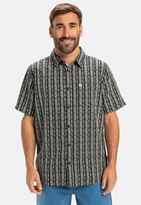 Camisa negra de manga corta con patrones florales blancos verticales y cierre de botones; cuenta con un pequeño logo en el lado izquierdo del pecho.