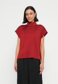 Punainen yläosa, jossa on korkea kaulus, lyhyet hihat ja väljä istuvuus. Valmistettu sileästä kankaasta, siinä on suora helma ja minimaalinen koristelu.