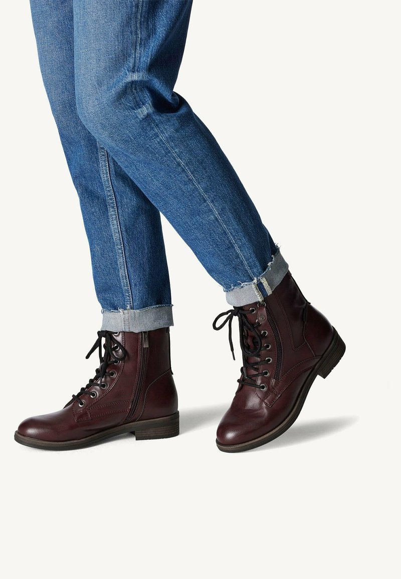 Botas de cuero burdeos con cordones, punta redonda, cremallera lateral y acentos texturizados. Llevadas con jeans de mezclilla azul arremangados.