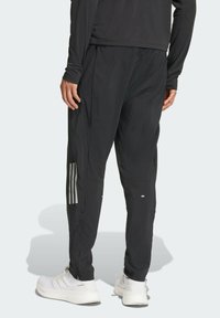 Sorte træningsbukser med en tætsiddende pasform, der har reflekterende striber, lynlåslommer og et let, struktureret stof. Hvide sko bæres.