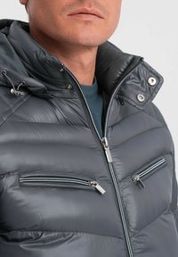 Giacca grigia imbottita con colletto alto, dotata di zip argentate su due tasche frontali e una texture liscia e lucida. Cappuccio regolabile con cordino.