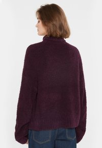 Übergroßer, flauschiger Pullover in tiefem Lila, mit hohem Kragen und lockeren Ärmeln, kombiniert mit Jeanshosen. Weiche Textur, lässige Passform.