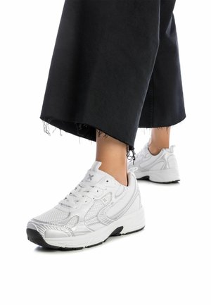 Zapatos deportivos blancos con un upper de malla texturizada, diseño de cordones y una suela de goma negra. Llevados con pantalones negros desgastados de longitud hasta el tobillo.