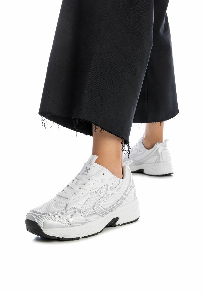 Zapatos deportivos blancos con un upper de malla texturizada, diseño de cordones y una suela de goma negra. Llevados con pantalones negros desgastados de longitud hasta el tobillo.