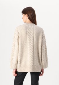 Beige neulepusero, jossa on teksturoitu kaapelikuosi, roikkuvat olkaimet ja ribattu hihansuu. Pehmeä, mukava materiaali, jossa on rento istuvuus.
