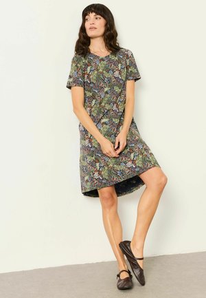 Frau lehnt an der Wand und trägt ein knielanges, kurzärmeliges Kleid mit grünen und braunen Blumenmustern sowie dunkelbraune Mary-Jane-Schuhe.