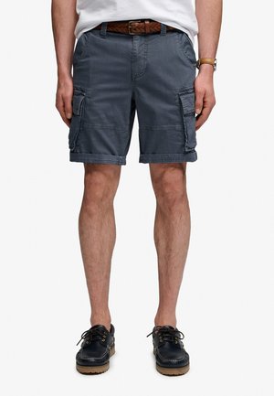 Mann trägt marineblaue Cargo-Shorts, geflochtenen braunen Gürtel, weißes Hemd, Uhr und schwarze Leder-Schnürschuhe.