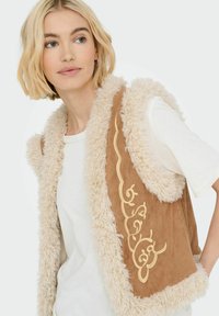 Braune Kunstsuede-Weste mit cremfarbiger, flauschiger Borte und kunstvollen goldenen Stickereien. Das Model trägt ein weißes T-Shirt darunter.
