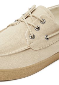 Sneaker in tela beige con occhielli in metallo, lacci rotondi e suola in gomma testurizzata. Presenta cuciture piatte e un design semplice e minimale.
