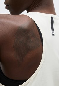 Κοντινό πλάνο του άνω μέρους της πλάτης ενός ατόμου με ένα σκούρο τατουάζ σε σχήμα φτερού, που φοράει λευκό αμάνικο τοπ με μαύρη λωρίδα λογότυπου.