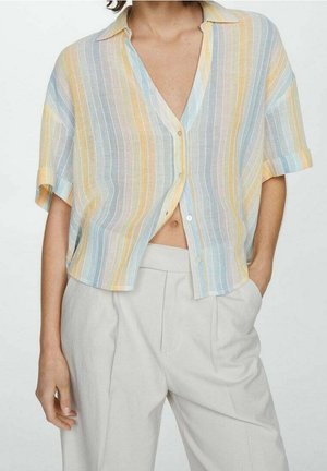 Chemise à manches courtes avec boutons et rayures pastel verticales, portée partiellement ouverte sur un pantalon plissé clair à taille haute.