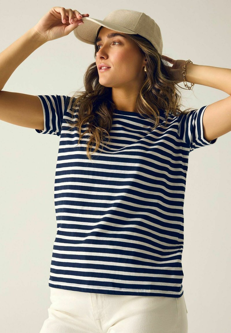 Frau, die eine beige Kappe richtet, trägt ein marineblau-weiß gestreiftes Shirt und weiße Hose, mit welligem Haar und einem goldenen Armband am Handgelenk.
