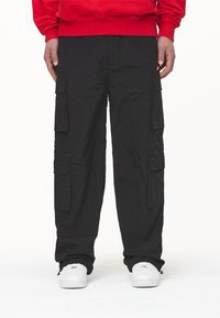 Pantaloni cargo negri cu multiple buzunare laterale, fabricați dintr-un material texturat. Asortați cu adidași albi și un hanorac roșu.
