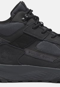 Scarpa sportiva nera caratterizzata da un mix di materiali texturizzati, pannelli in rete e suola in gomma con un motivo esagonale. Gli accenti includono un logo sul lato.