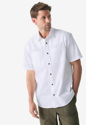 Chemise blanche à manches courtes avec une finition texturée, deux poches poitrine et des boutons noirs. Portée avec un pantalon vert olive.