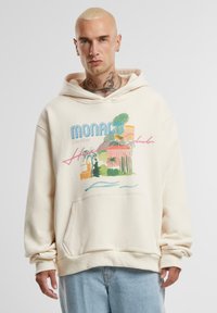 Krémová mikina s kapucí, kterou zdobí grafický tisk Monaka, palmami a stylizovaným textem. Obsahuje přední kapsu a nastavitelné šňůrky na kapuci.