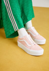 Pink suede sneakers with white accents and thick white soles, paired with green patterned pants featuring pink side stripes.