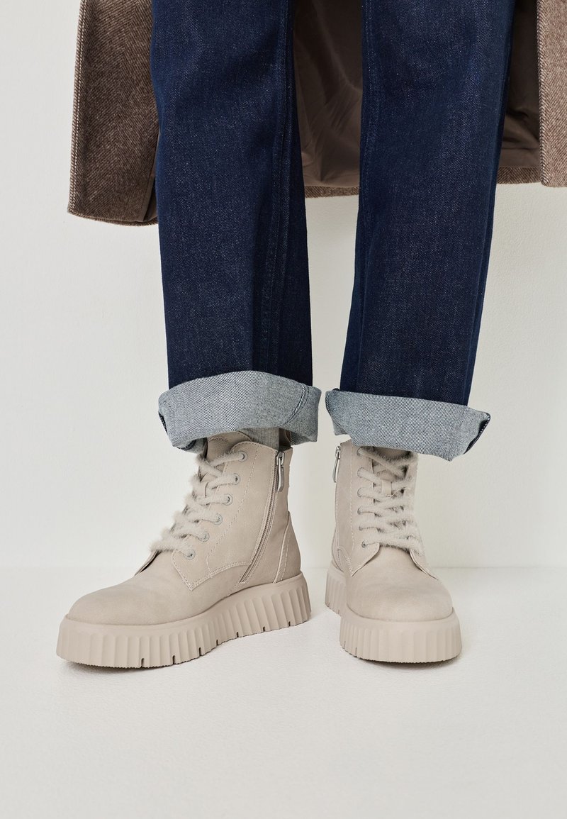 Beige Wildleder-Stiefeletten mit einer dicken, geriffelten Sohle, ausgestattet mit einem seitlichen Reißverschluss und einer schnürbaren Vorderseite. Getragen mit umgeschlagenen dunkelblauen Jeans.