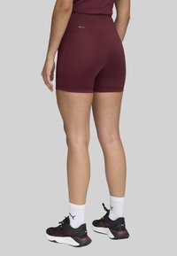 Burgundy athletic shorts with a high waistband, made of stretchy fabric. Features a small logo detail at the back. Paired with black sneakers and white socks.