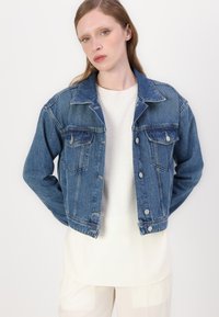 Denim-jakk, millel on klassikaline sinine pesu, nööbid ees, kaks rinna taskut ja kohandatud lõige, kihtide kaupa heledat särki.