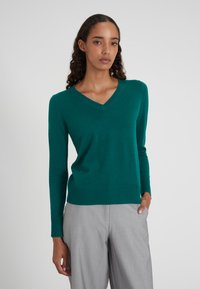 Femme portant un pull V en teal et un pantalon gris clair, se tenant avec une main dans la poche devant un fond uni.