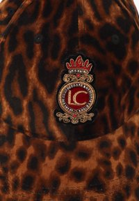 Cappello in velluto a stampa leopardata marrone con un emblema ricamato con una corona e "LC" in rosso, circondato da accenti decorativi.