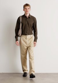 Camisa marrón de botones, pantalones beige y zapatos negros. El modelo lleva un bolso de hombro gris. Diseño simple de pierna recta.