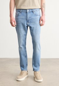 Világoskék farmernadrág, slim fit szabással, öt zsebbel és diszkrét kopásokkal. Bézs sportcipővel párosítva a semleges megjelenés érdekében.