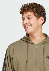 Sweat-shirt zippé vert olive en tissu doux, avec une finition texturée, des cordons de serrage et des bandes noires sur les manches.