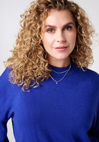 Koningsblauwe trui met een hoge hals, gemaakt van zachte stof. Draagt meerdere zilveren kettingen in laagjes. Krullend blond haar omlijst het gezicht.