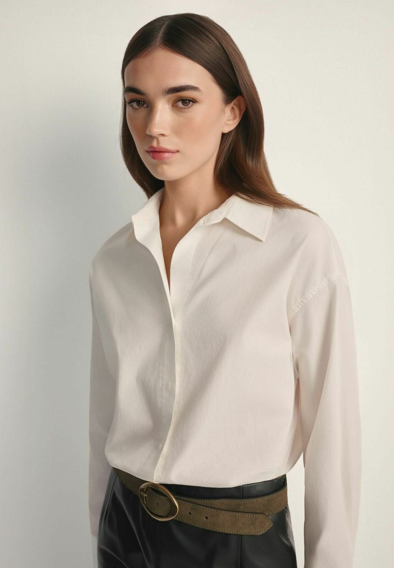 Młoda kobieta o długich brązowych włosach ubrana w białą koszulę zapinaną na guziki, wpuszczoną w czarne spodnie z brązowym paskiem, na jednolitym tle.