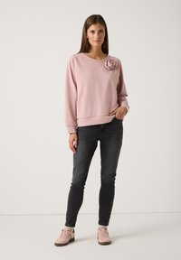 Sweatshirt rose clair avec un embellissement en 3D sur l'épaule gauche, associé à un jean skinny gris foncé et des baskets rose clair.